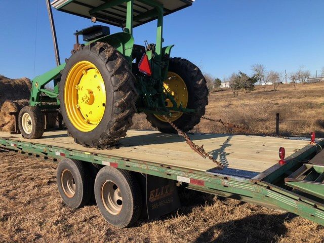 John Deere tractor on a flatbed trailer, secured with chains. Green and yellow colors, outdoor setting.