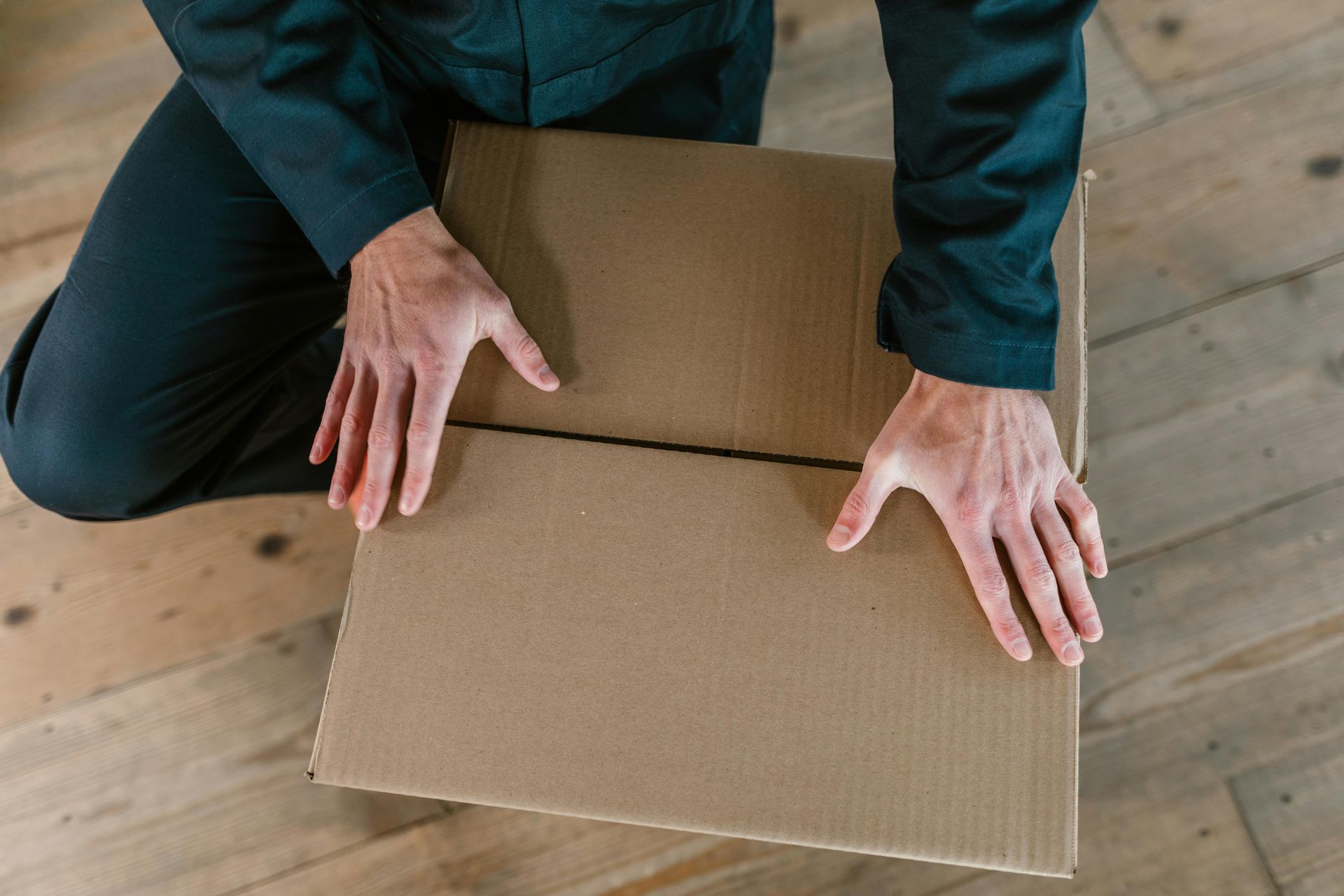 Person kneeling, opening a cardboard box on a wooden floor, hands visible.