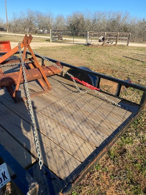 A rusty farm implement is secured on a wooden-floored trailer with chains, outdoors.