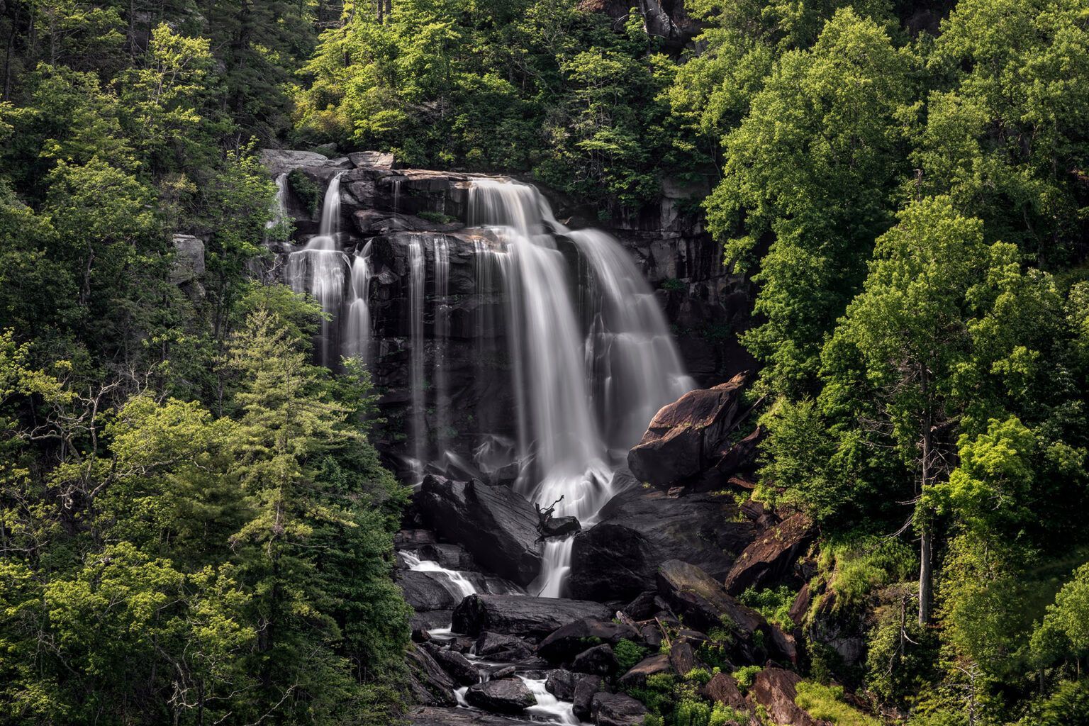 Waterfall cascading down dark rocks, surrounded by lush green trees.