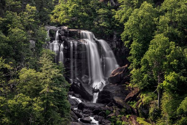 Waterfall cascading down dark rocks, surrounded by lush green trees.