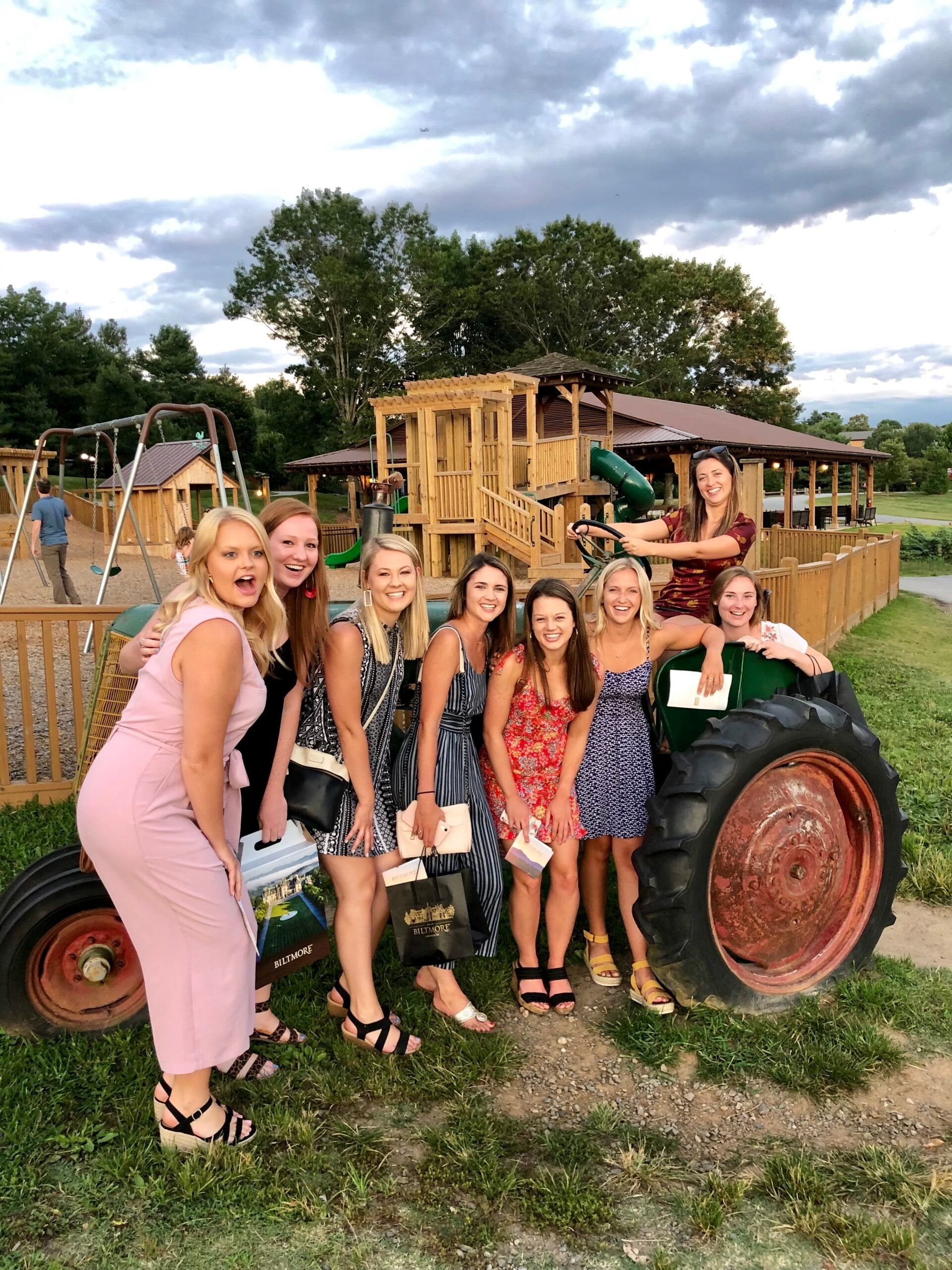 Group of women posing on a tractor outside, smiling. Background includes a playground and trees.