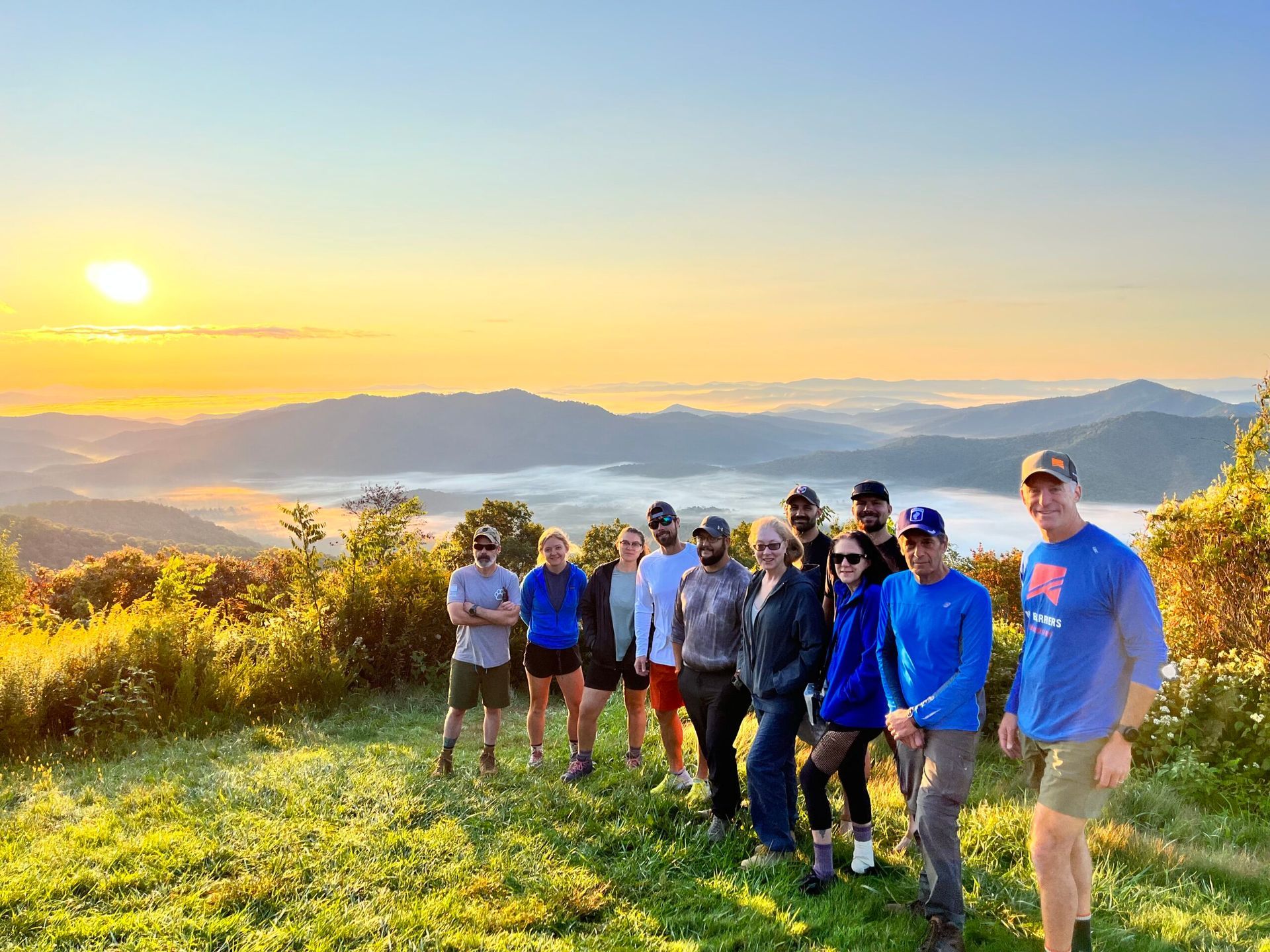 Group of people stand on a mountain at sunrise. Golden light shines over a valley and peaks.