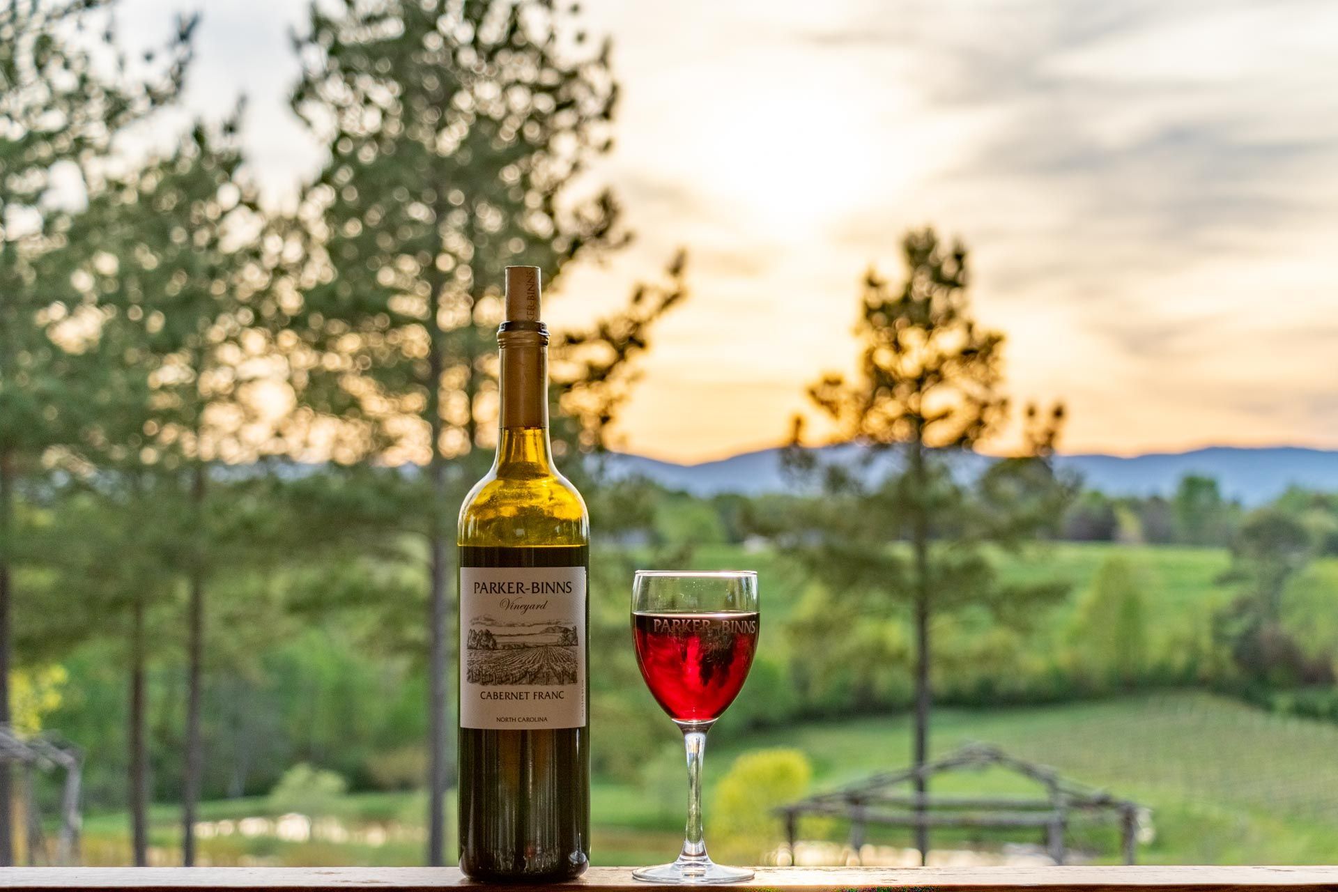 Parker Bins bottle of red wine and a glass filled with red wine on a wooden railing against the vineyard landscape at sunset.