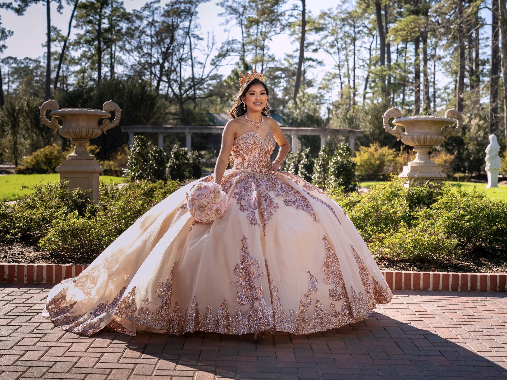 Woman in a pink embellished ball gown standing in a garden courtyard with stone urns and trees.
