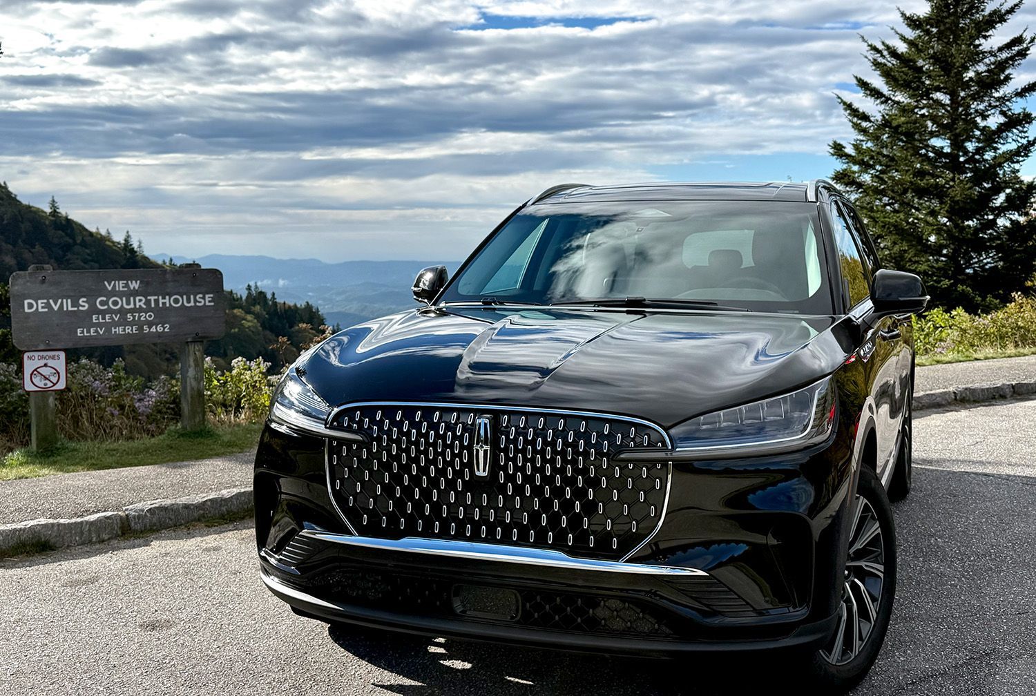 Black Lincoln Aviator SUV parked at Devil's Courthouse scenic overlook off the Blue Ridge Parkway. Mountains in the background, cloudy sky.