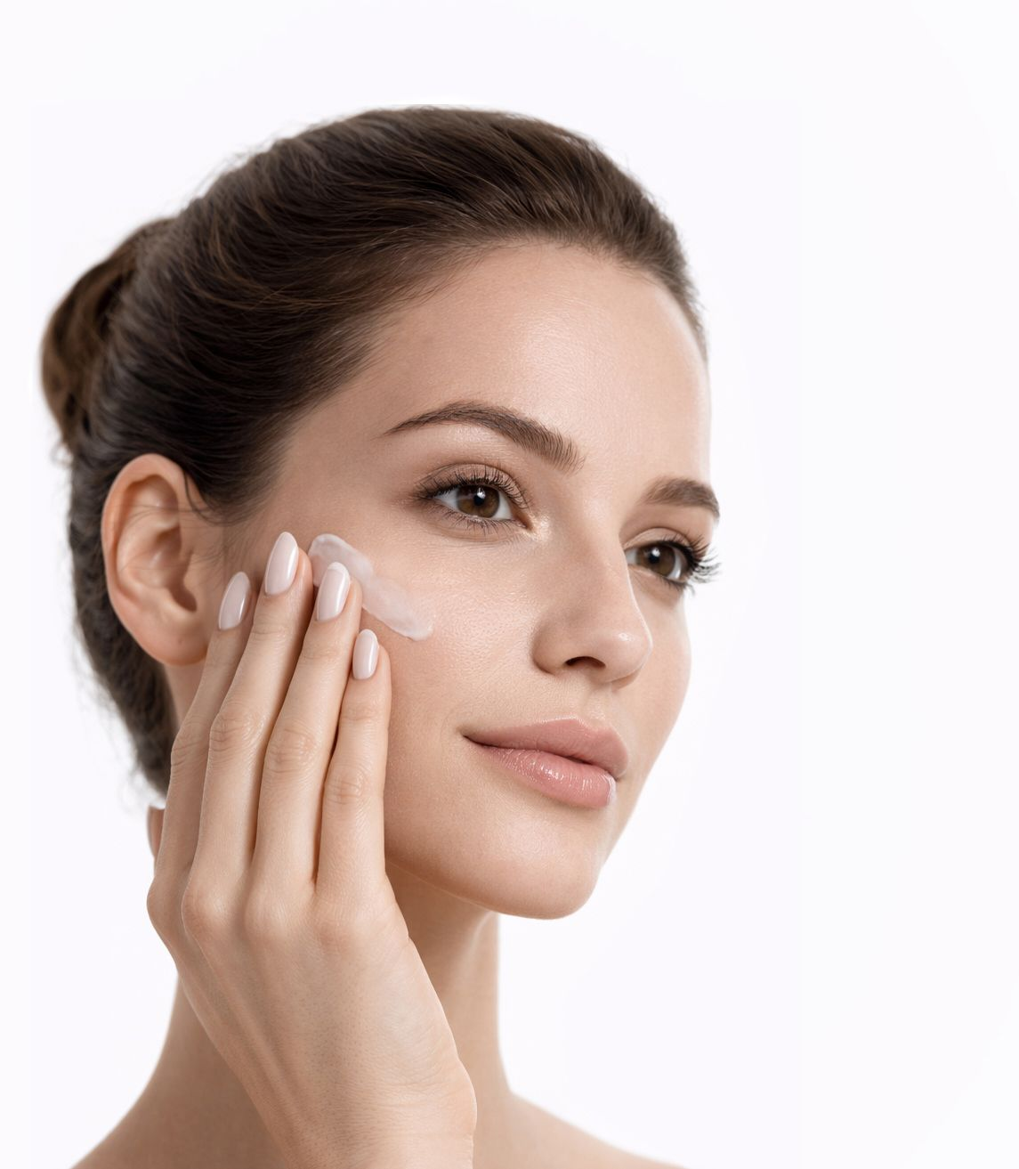 Mujer aplicándose crema en la mejilla. Primer plano, piel clara, cabello castaño recogido, fondo blanco.