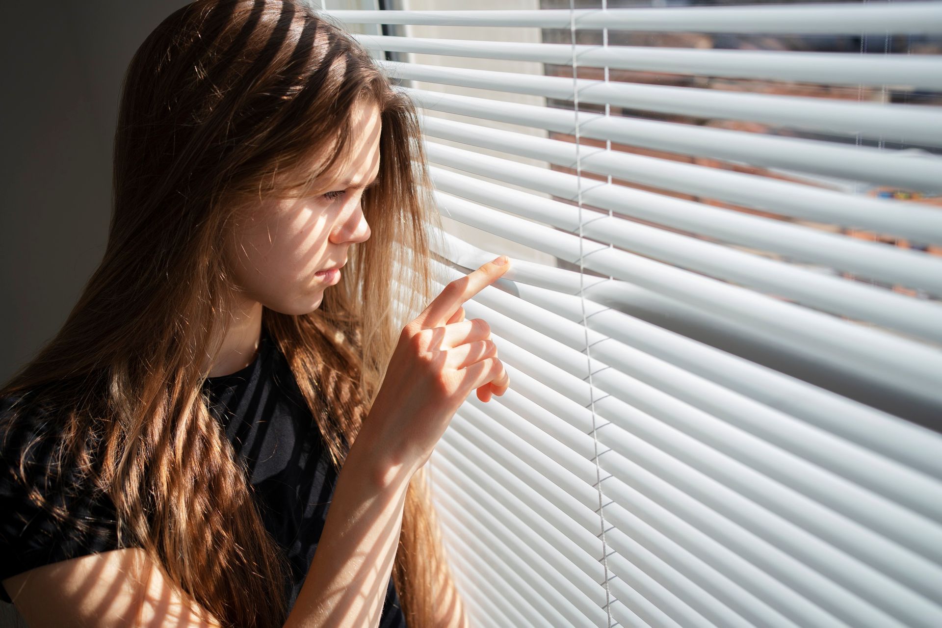 A young woman is looking out of a window through blinds.