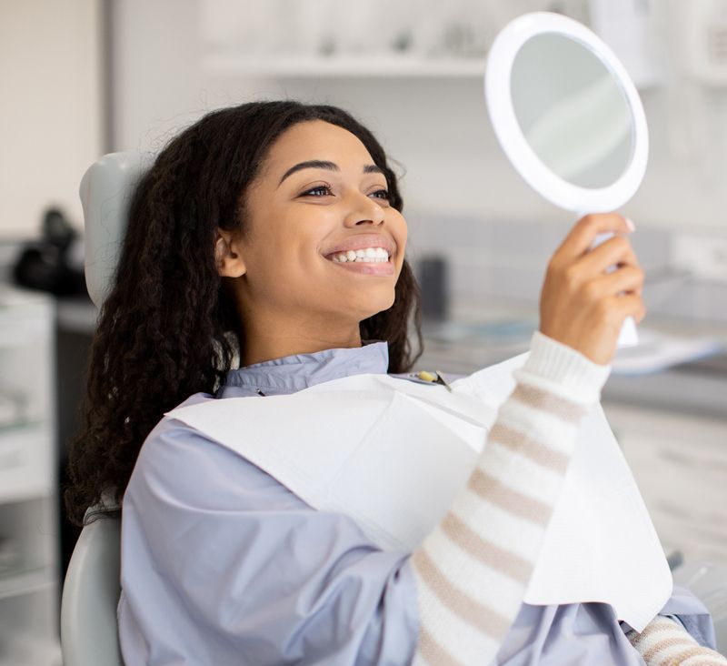 A woman is sitting in a dental chair looking at her teeth in a mirror.