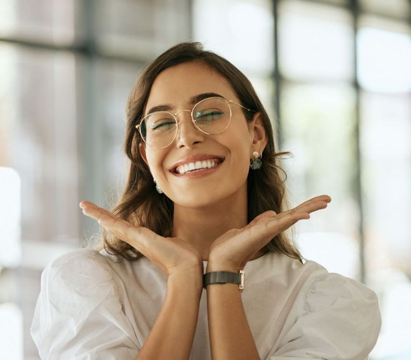 A woman wearing glasses is smiling with her hands on her face.