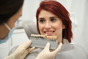 Woman in a dentist chair smiles at her new teeth, holding a mirror, with a masked assistant in the background.