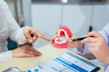 Woman in a dentist chair smiles at her new teeth, holding a mirror, with a masked assistant in the background.
