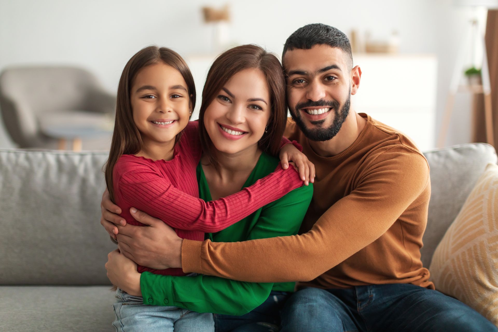 Smiling family of three hugging on a gray couch in a bright living room