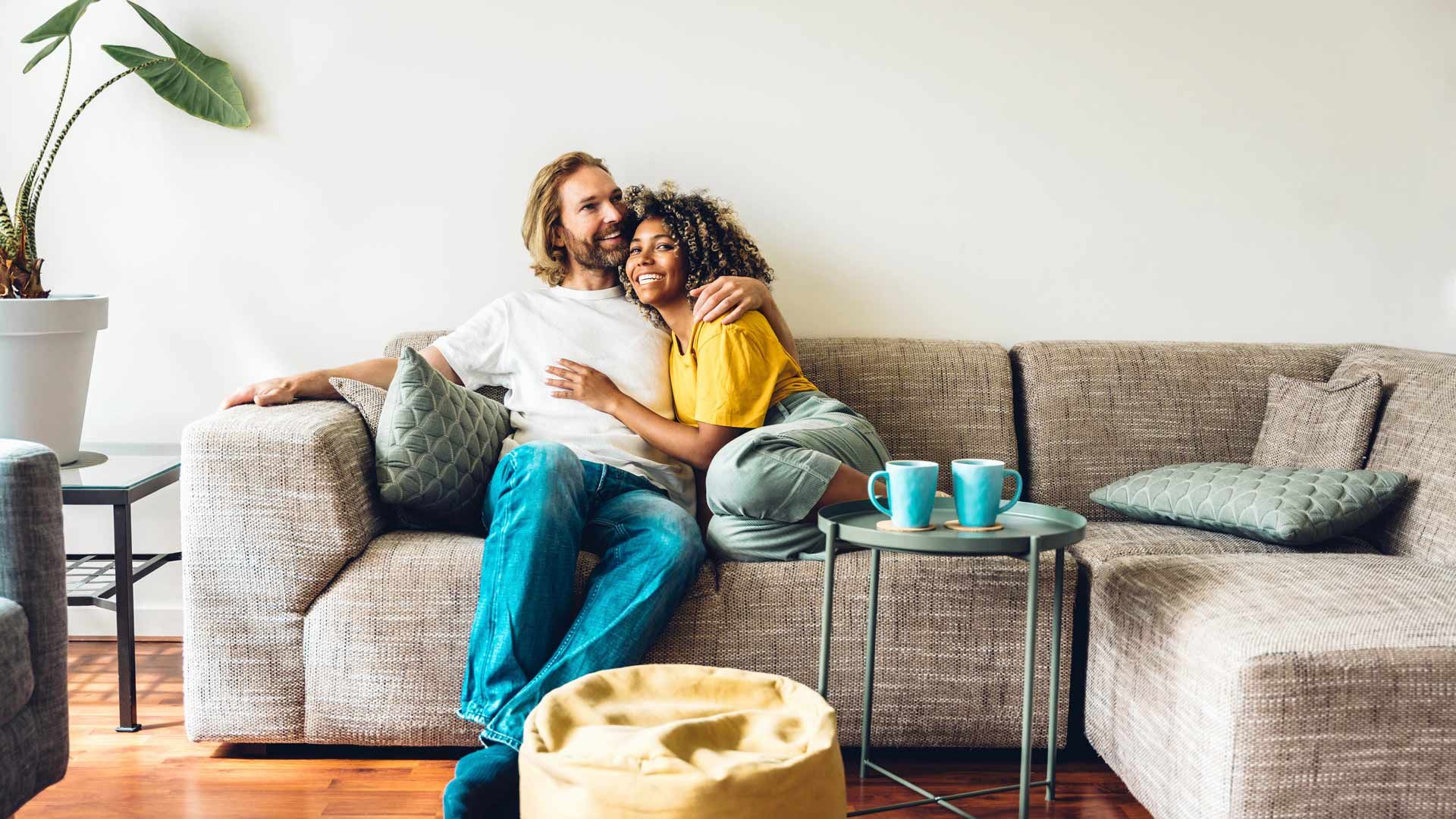 Two people cuddling on a beige couch in a bright living room with teal pillows and a small table. Two people cuddling on a beige couch in a bright living room with teal pillows and a small table.