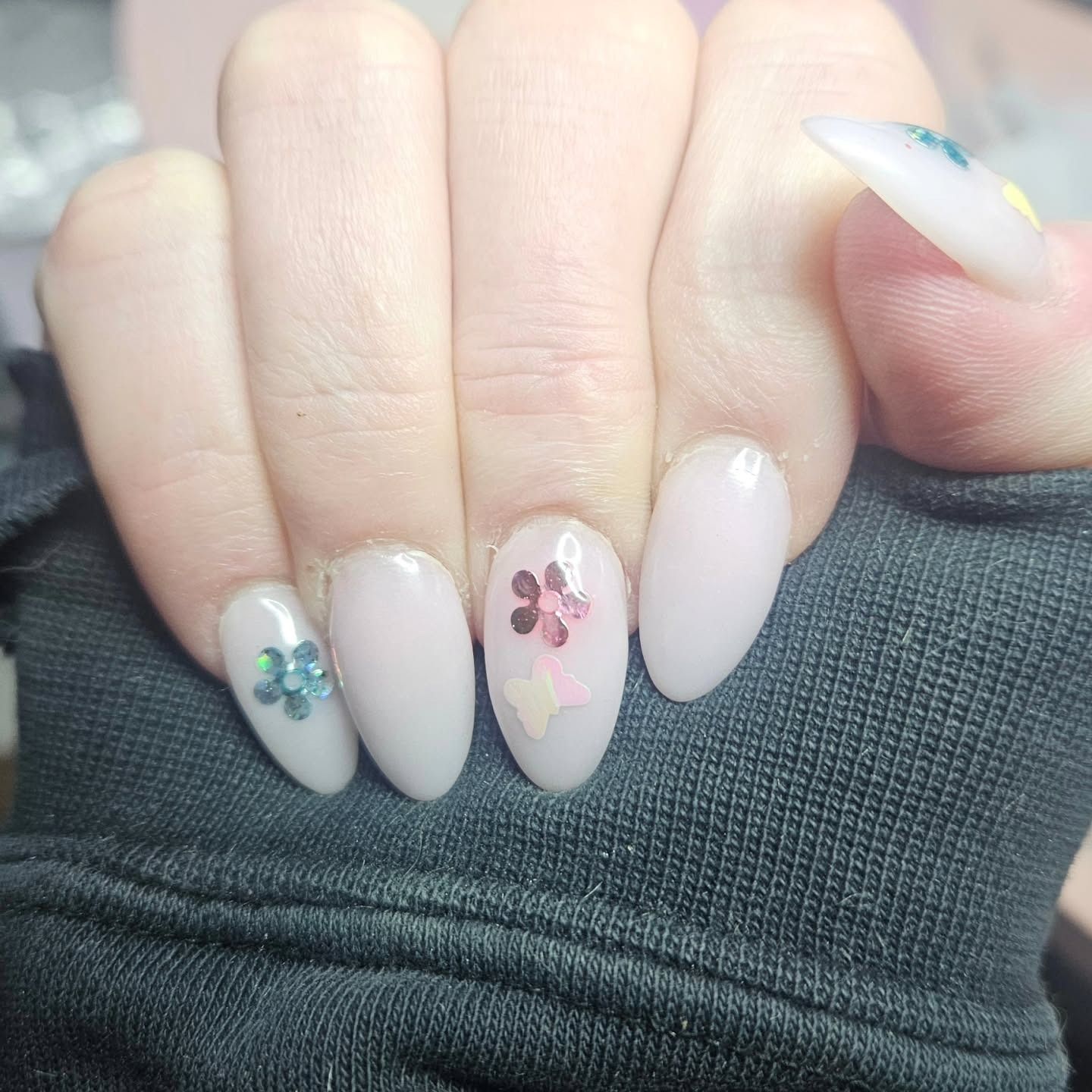 A hand with almond-shaped, milky white nails. Some nails feature small, colorful floral accents and gem embellishments.