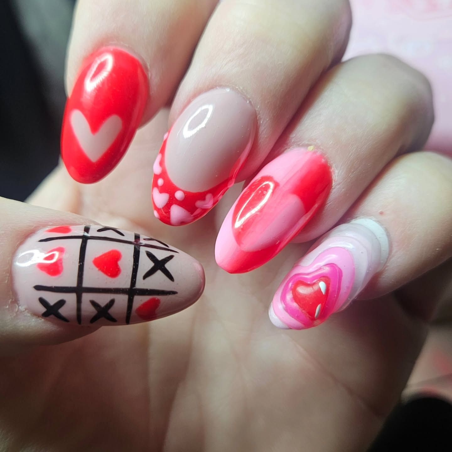 Oval-shaped nails with festive Valentine's Day nail art, featuring hearts, a tic-tac-toe grid, and pink and red designs.