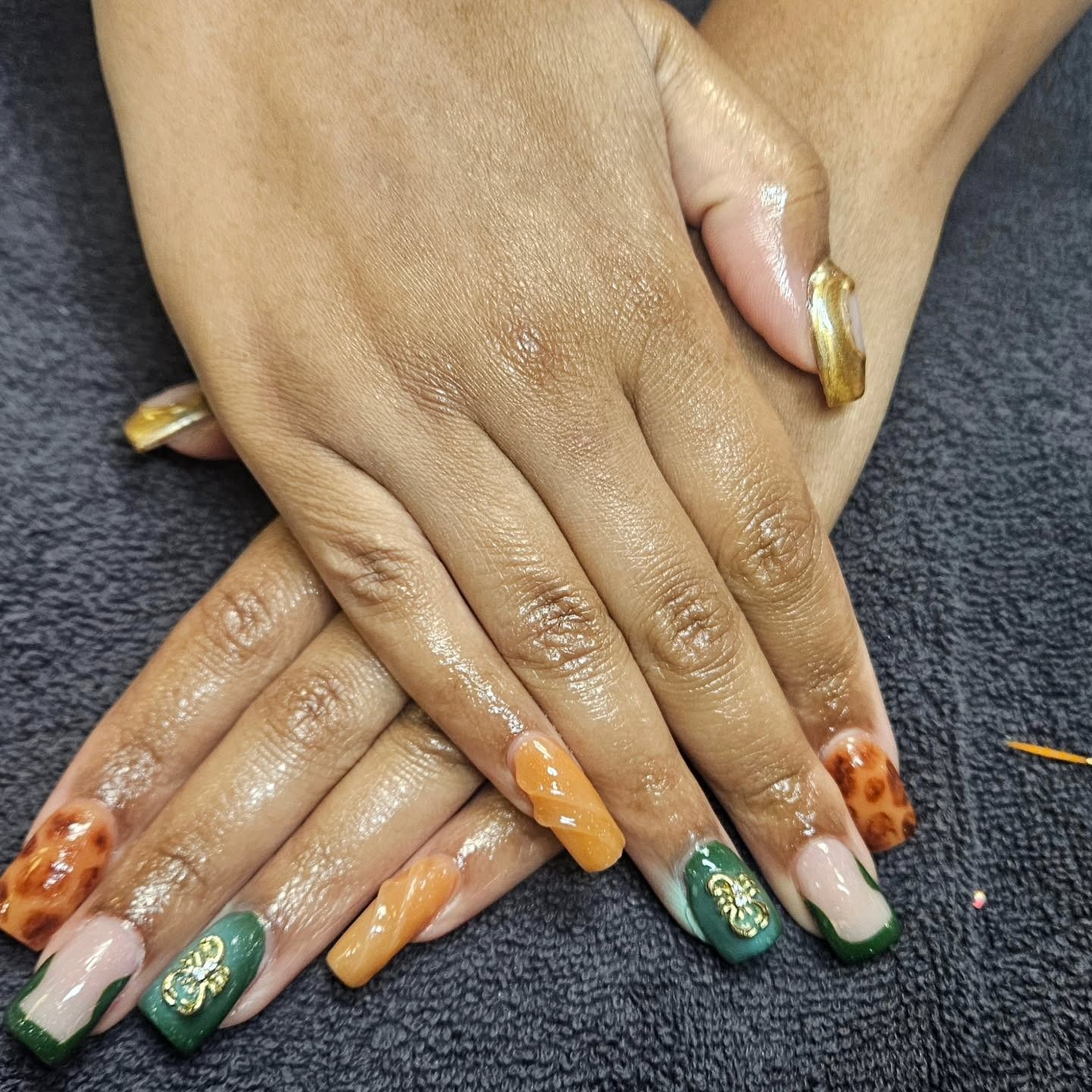 A close-up of hands with long, manicured nails featuring green, orange, and leopard print designs with gold accents.