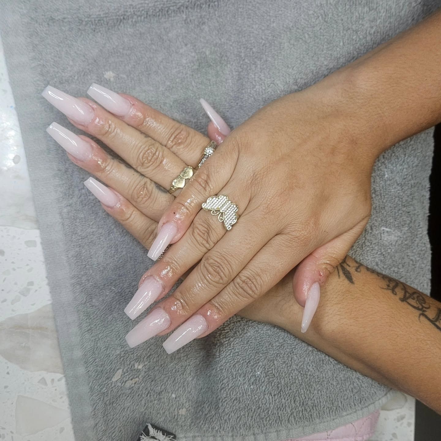 Two hands with long, light pink coffin-shaped nails and butterfly rings, resting on a gray towel.