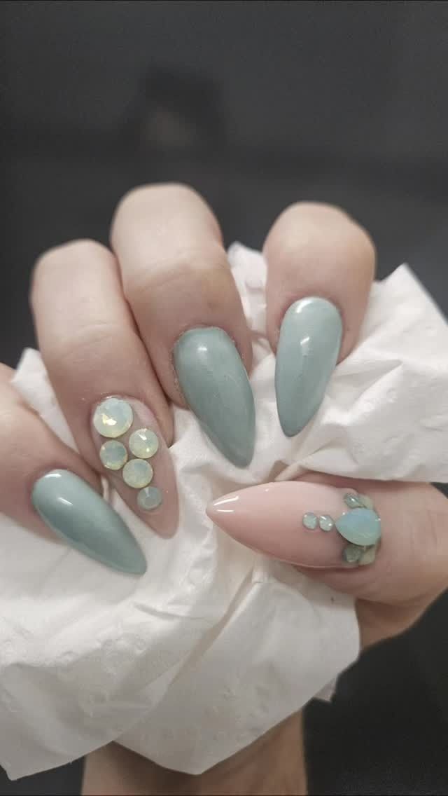 A hand with almond-shaped nails features light blue and pale pink polish, accented with clusters of iridescent gems.