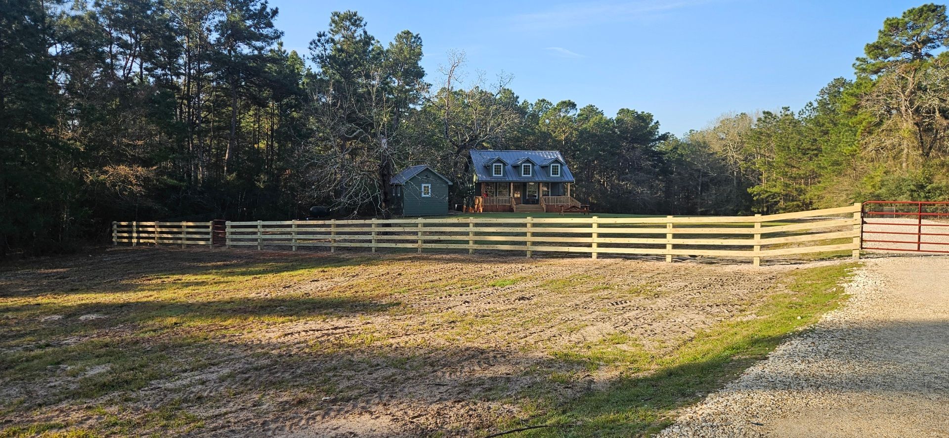 Wooden fence surrounding a field with trees and a house in the background. Sunny day.