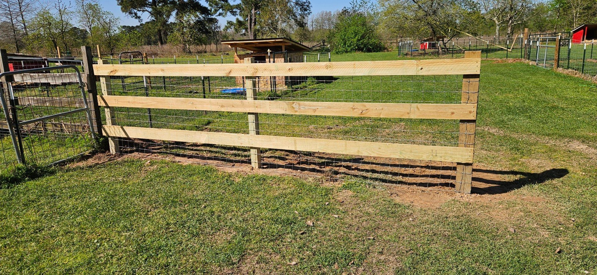 Wooden fence in a grassy field with a shed and trees in the background under a blue sky.