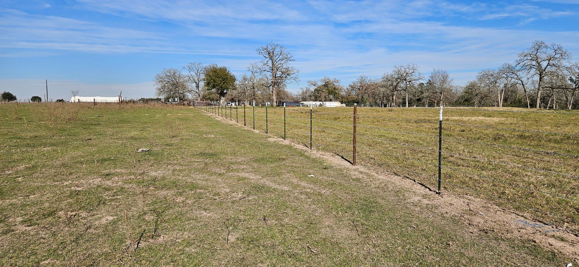 A grassy field with a fence in the foreground under a blue sky. Bare trees are visible in the distance.