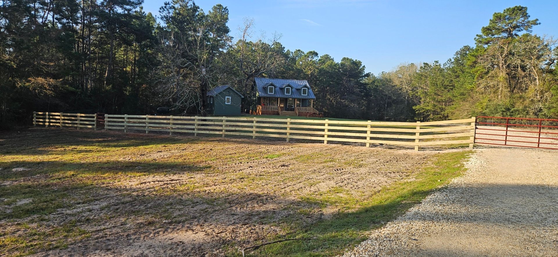 Wooden fence surrounds a grassy field, with a small cabin and trees in the background under a blue sky.