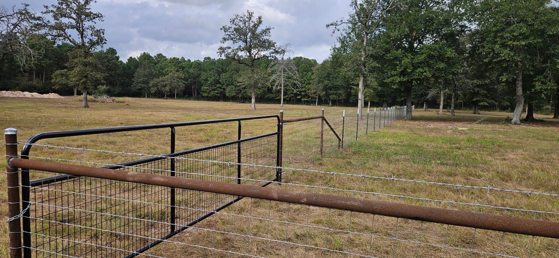 A rusty gate in a grassy field, with trees in the background under a cloudy sky.