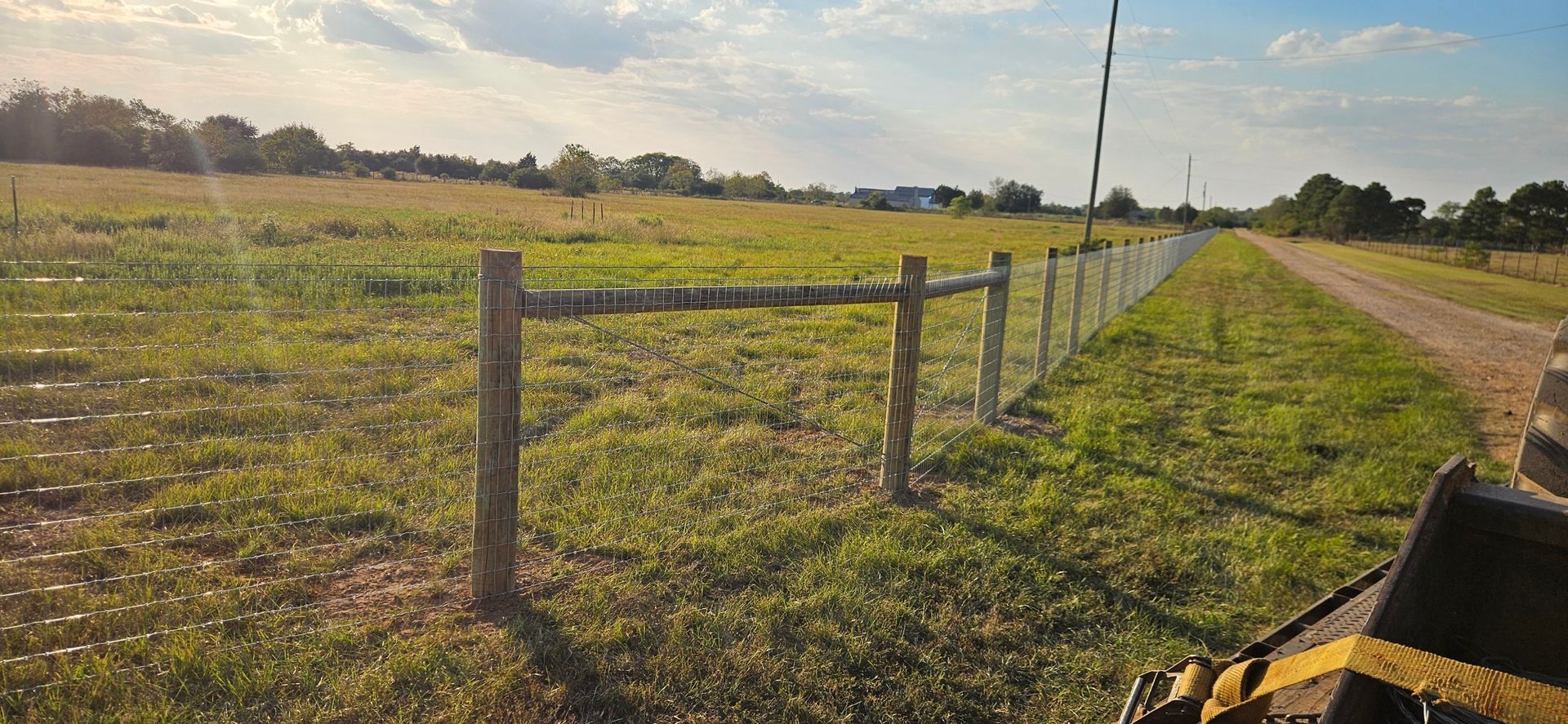 A wooden fence runs along a grassy field beside a dirt road under a sunny sky.