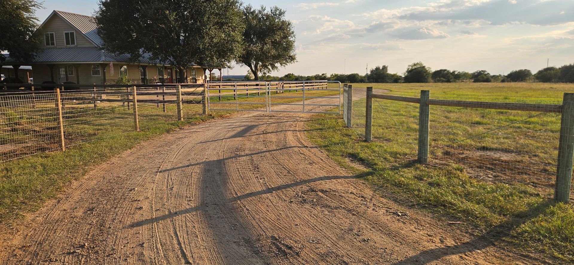 Dirt road leading to a yellow house with a gray roof and a wooden fence along a grassy field.