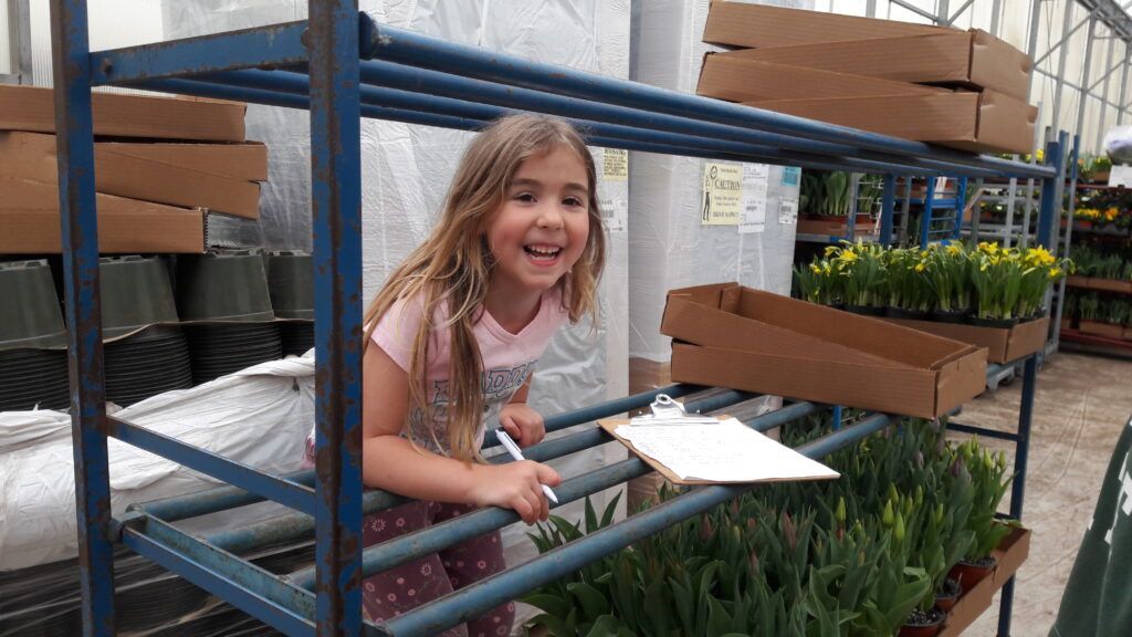 A little girl is standing in a greenhouse holding a clipboard.