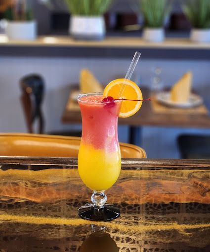 A pink and yellow cocktail with an orange slice and a cherry on a bar.