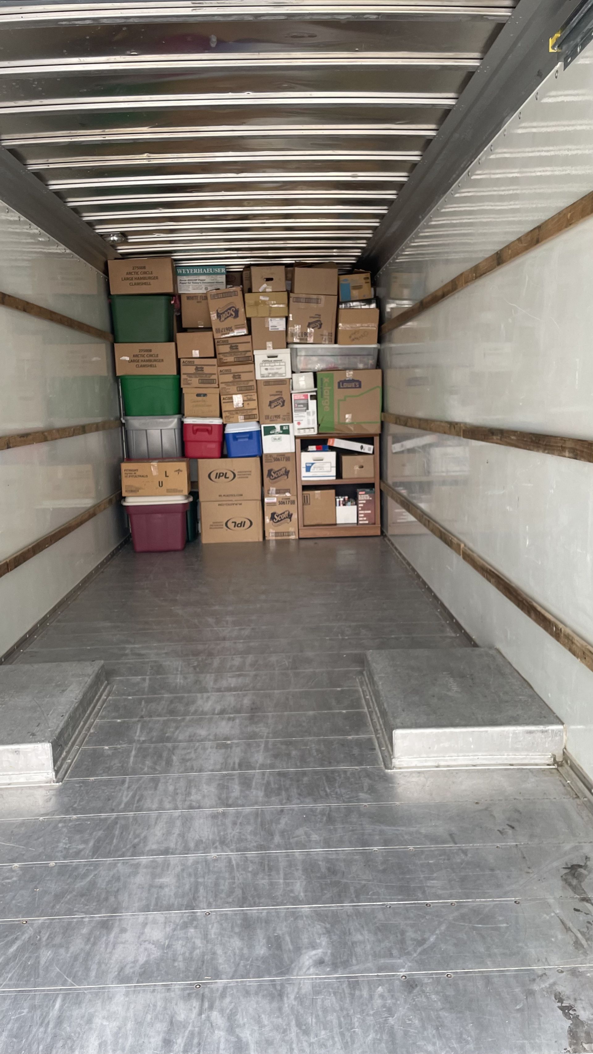 Inside of a moving truck with boxes piled high against the back wall; gray floor and white walls.