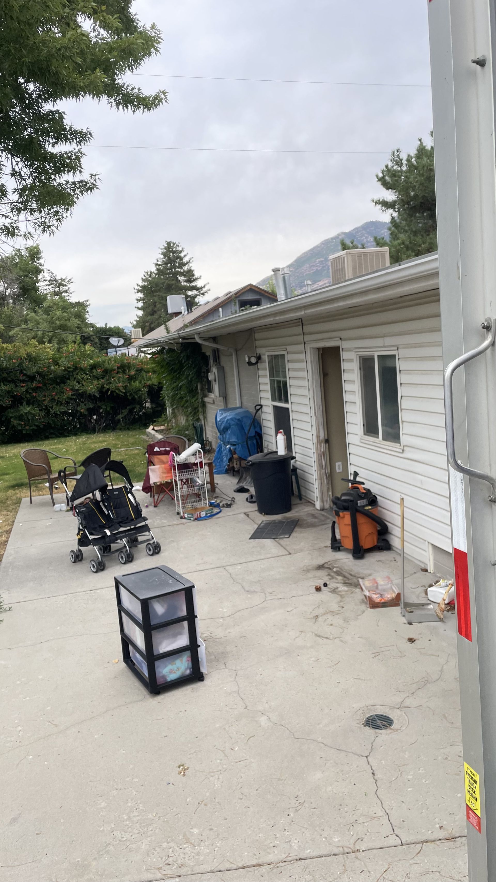 Exterior of a white building with clutter on a concrete patio. Mountains in the background, cloudy sky.