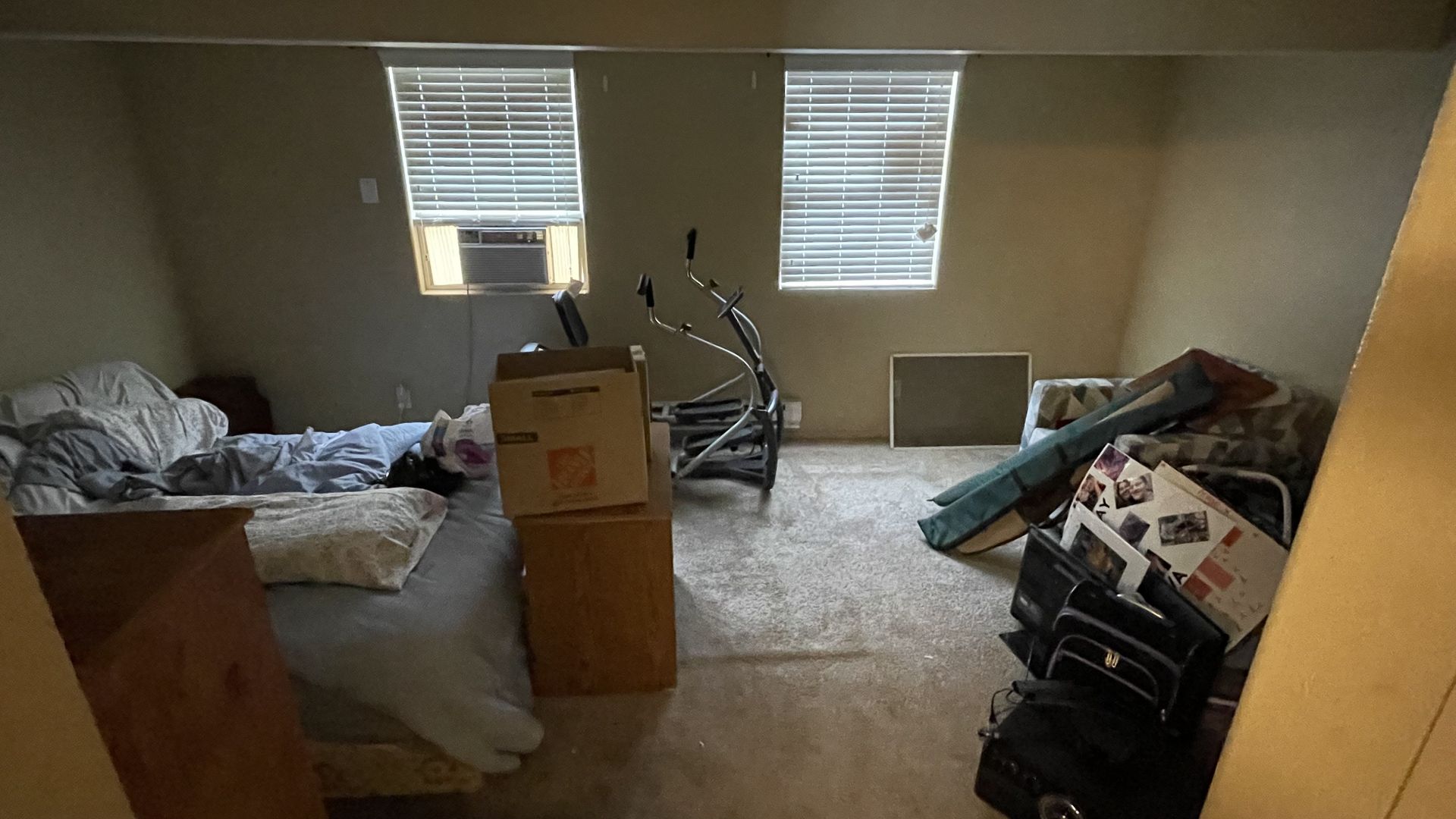 Cluttered room with two windows, a bed, cardboard boxes, and exercise equipment. Beige walls and carpet.