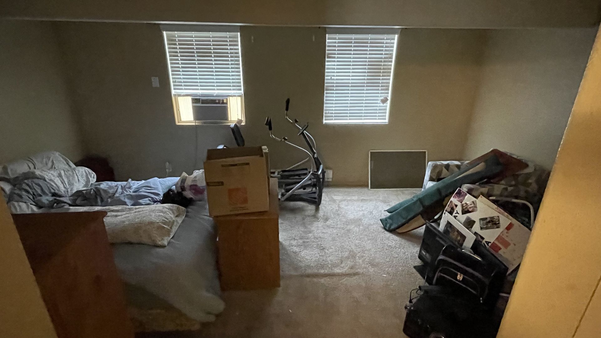 A cluttered room with two windows, boxes, a crib, and a bed with various items.