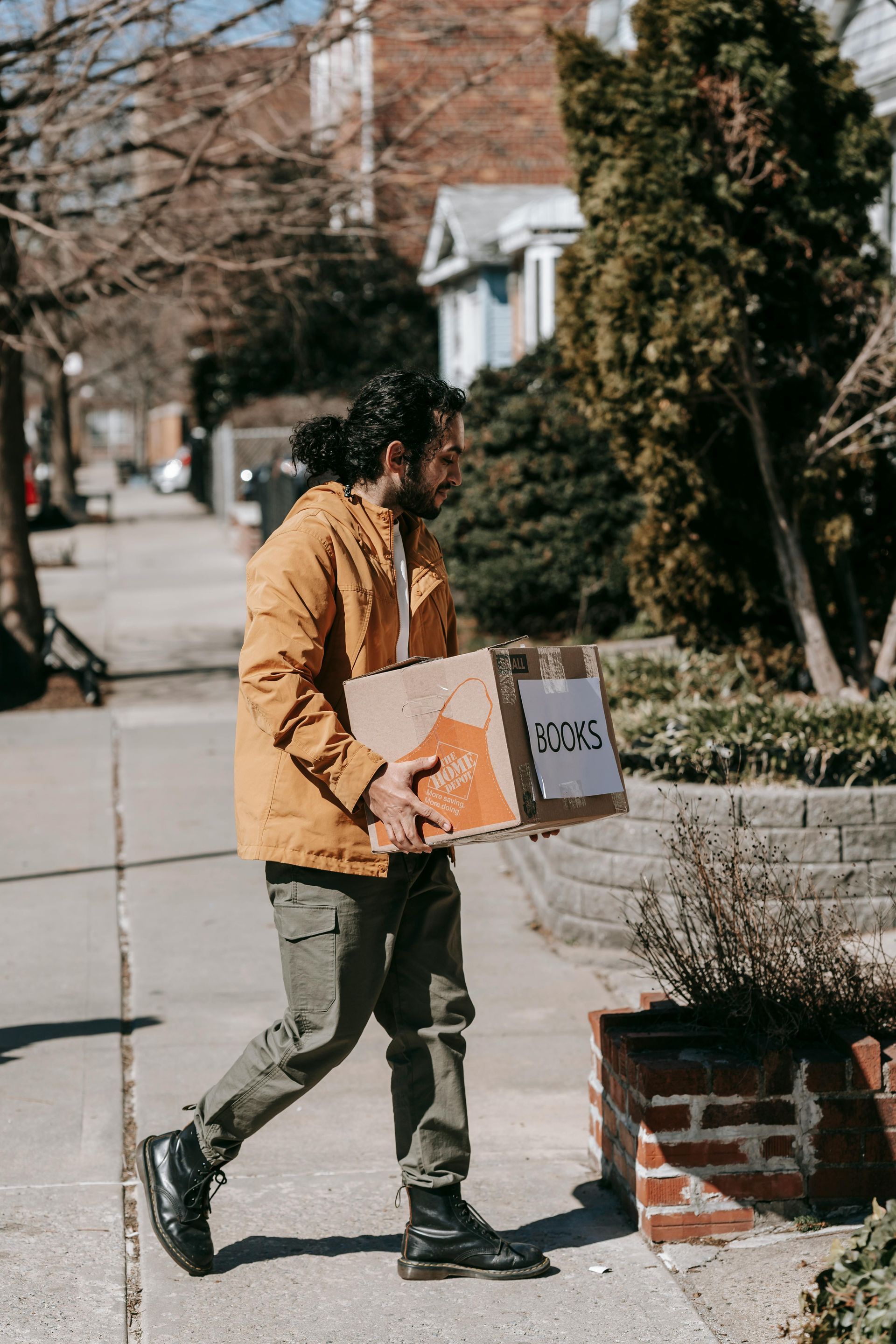 Man carrying a cardboard box on a sidewalk in front of a house. He wears a yellow jacket, green pants and black boots.