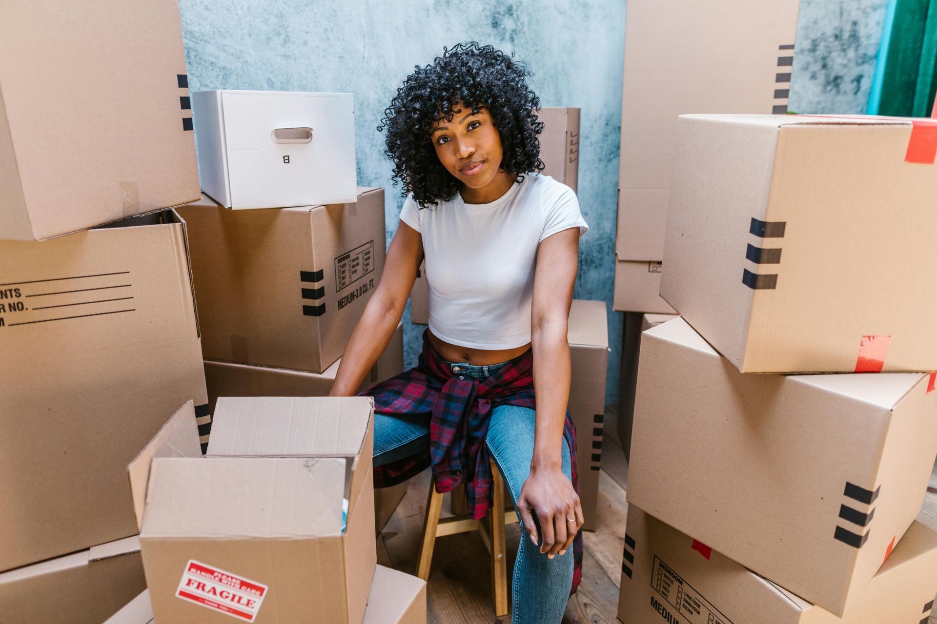 Woman sitting on a stool among moving boxes; wearing a white shirt and jeans, looking at camera.