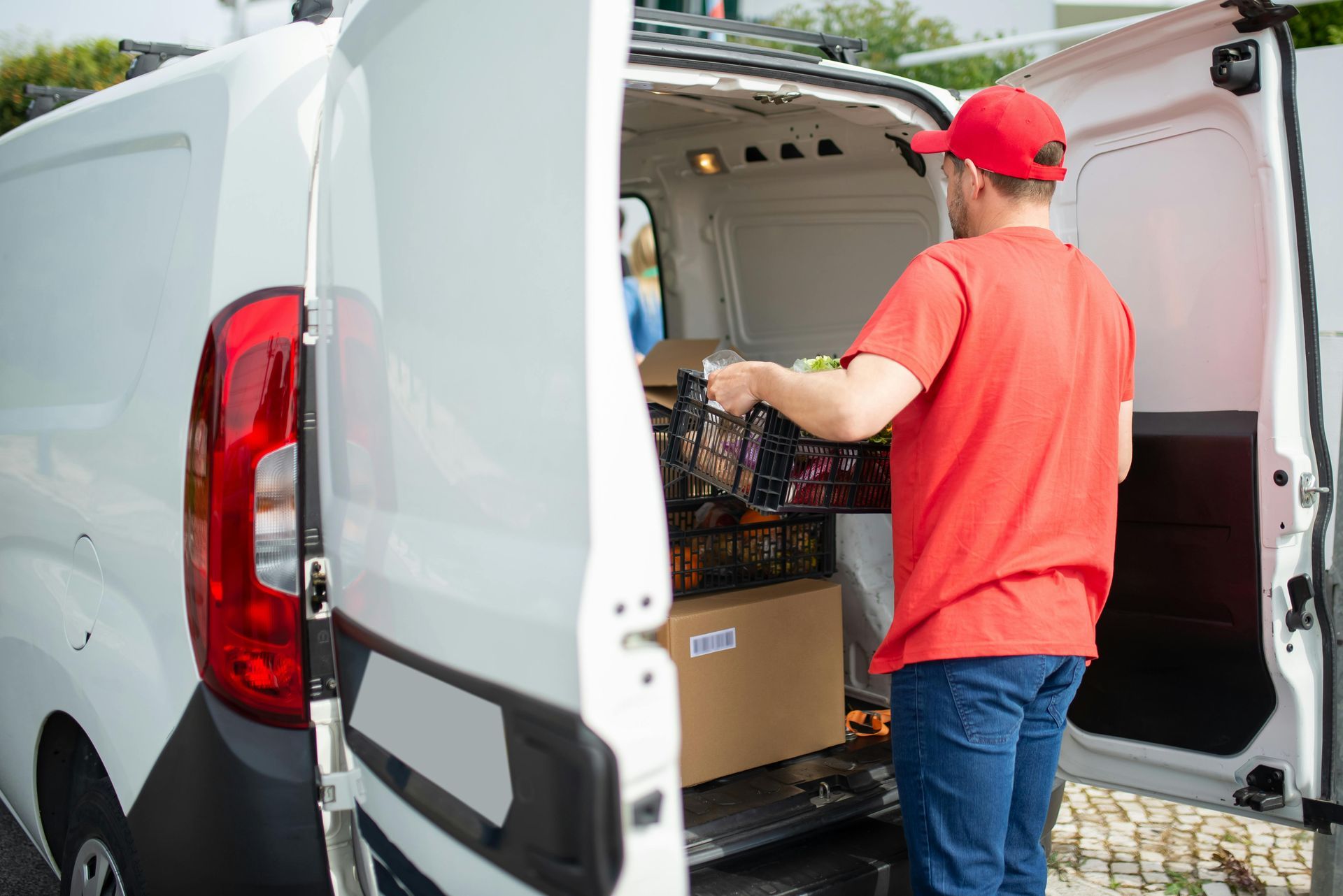 Delivery person loading groceries into a white van; red shirt, cap.
