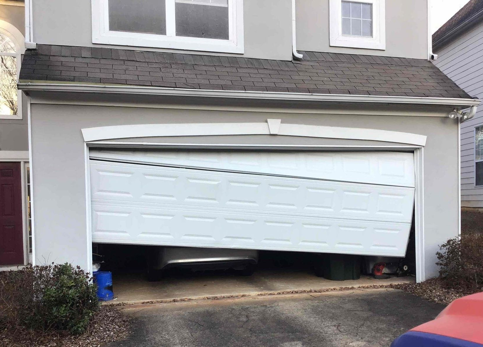 Garage door partially open, appearing damaged. Car inside. White door, gray stucco house, dark roof.