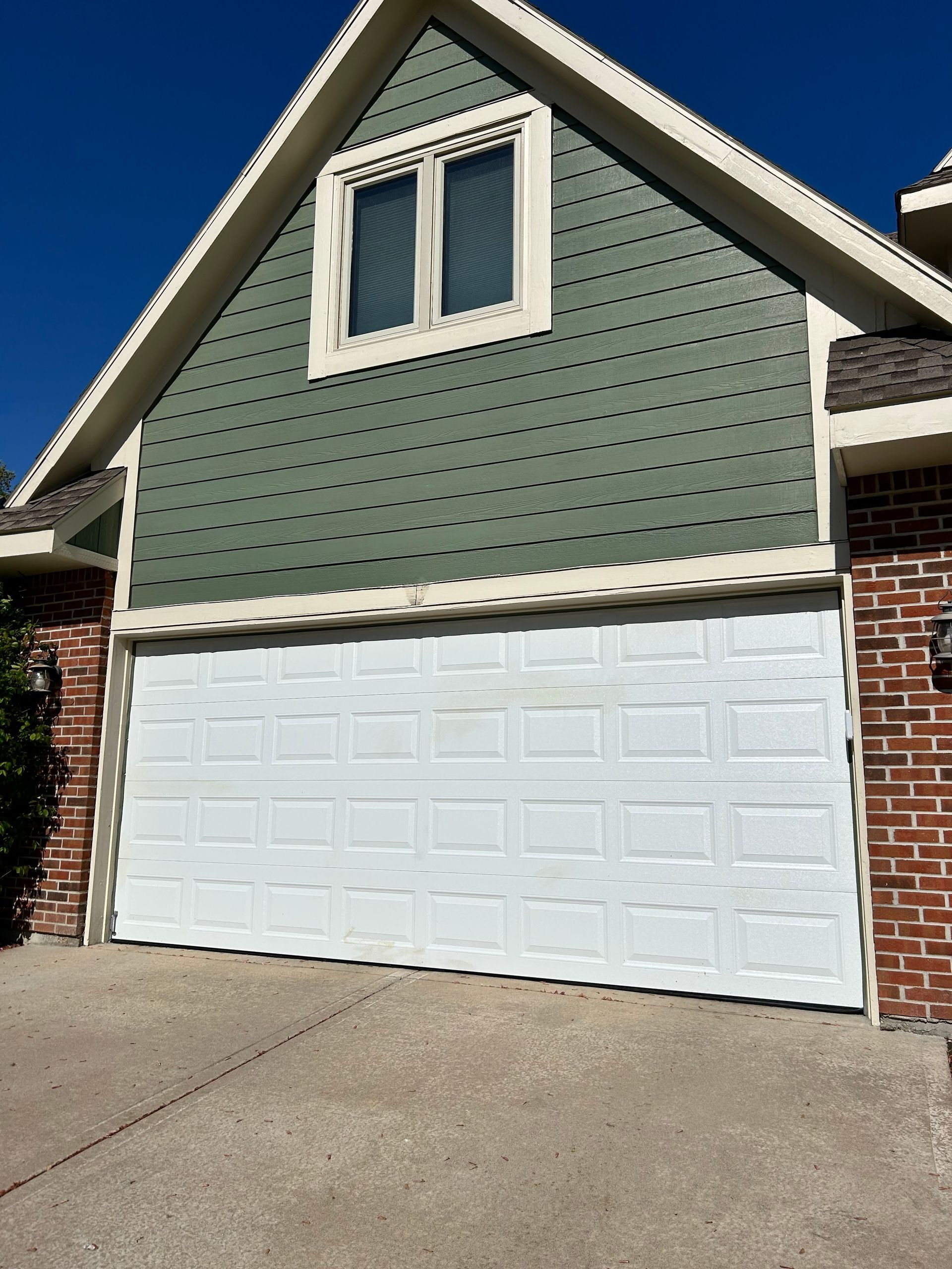 White garage door beneath a green-paneled gable roof with a window, set against a brick wall.