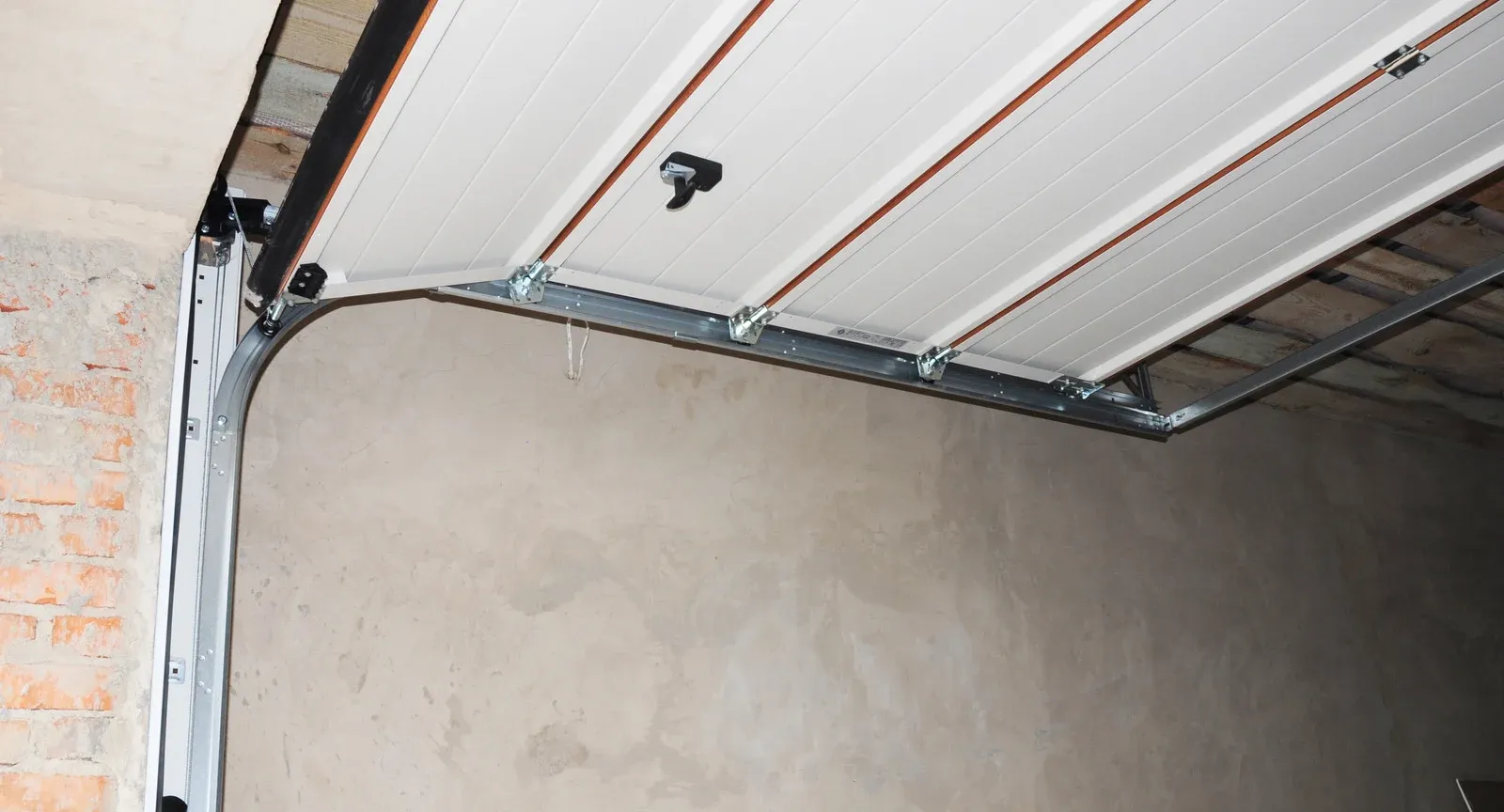 Interior view of a garage door, partially opened, with white panels and metal tracks against a brick and plaster wall.