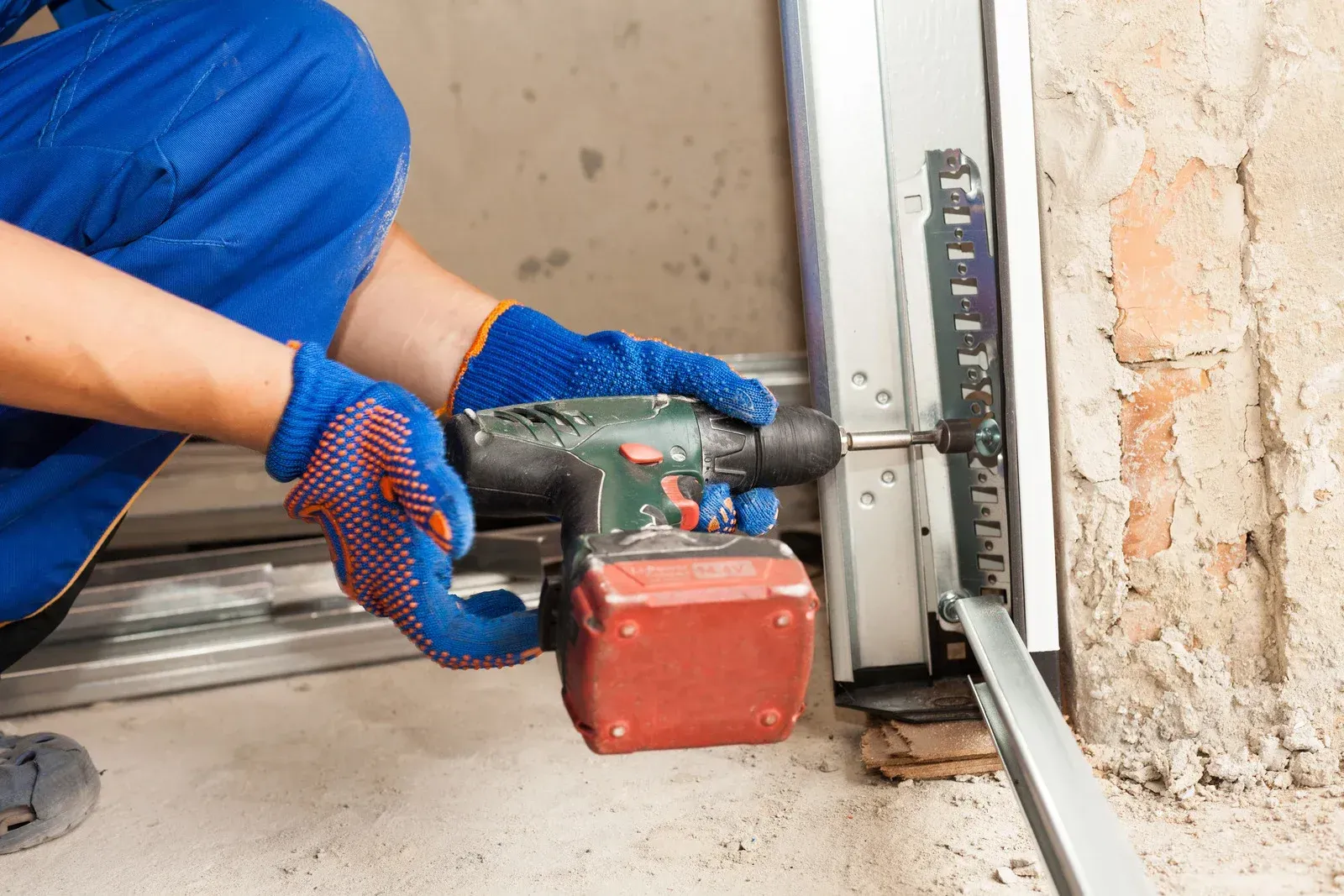 Person in blue overalls and gloves using a drill to install a metal garage door component near a brick wall.