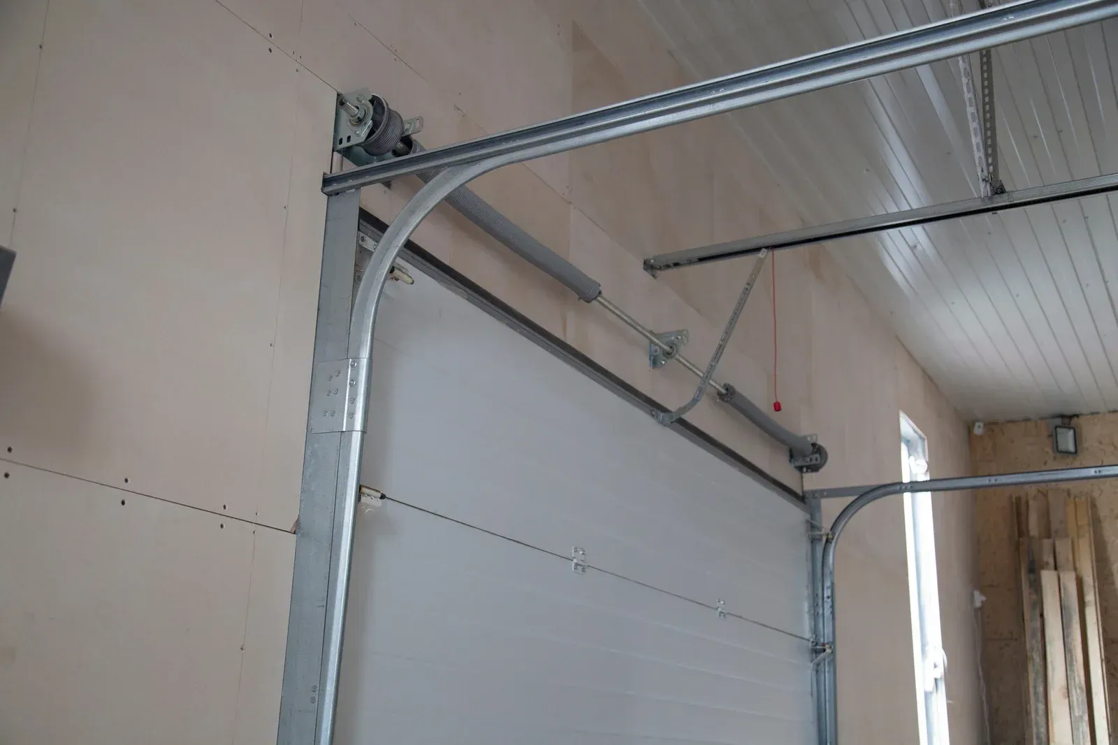 Garage door partially open, white door panel, silver tracks and hardware attached to tan wall and ceiling.