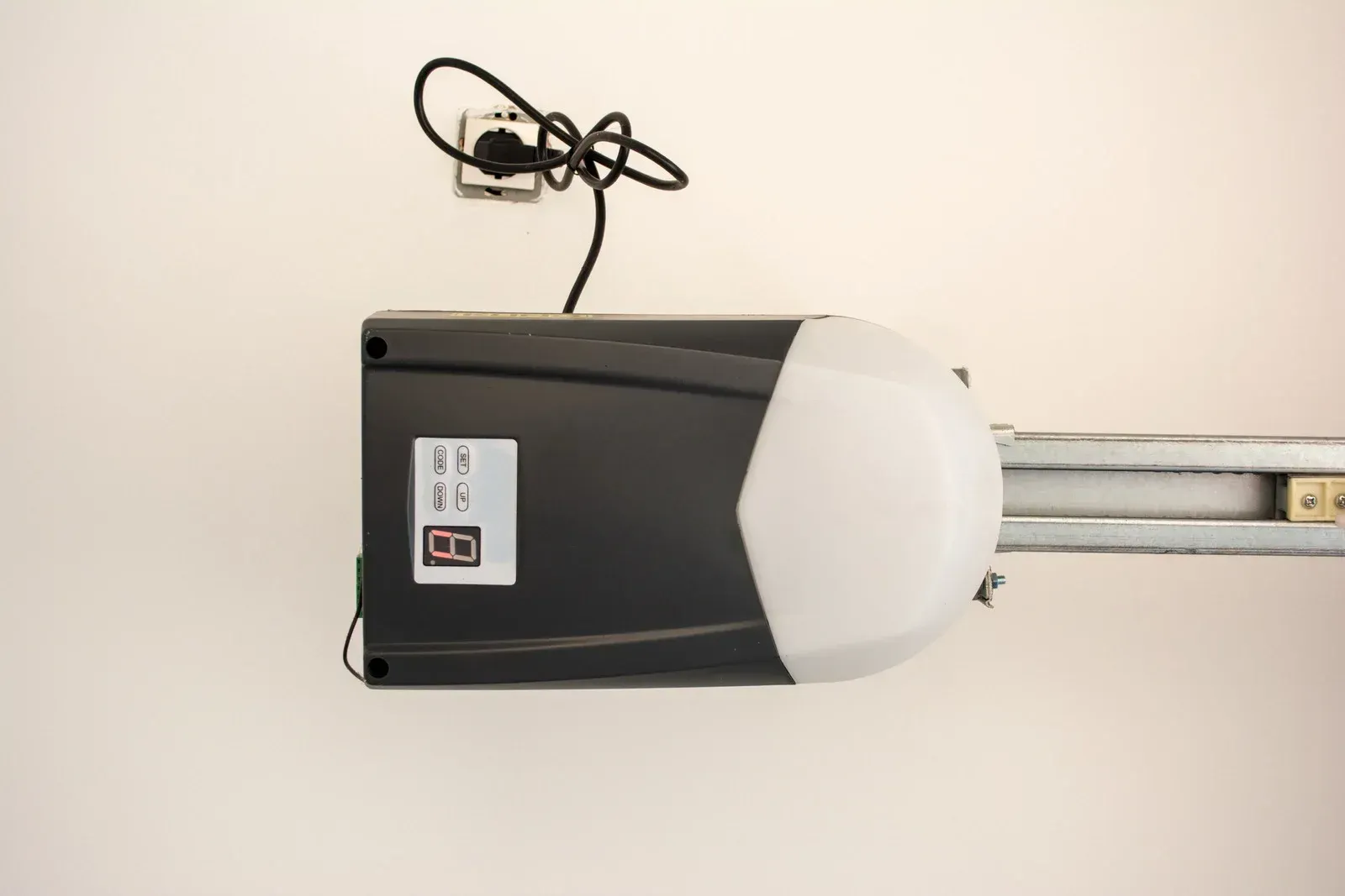 Garage door opener mounted on a beige wall, with a cord plugged into a nearby outlet.