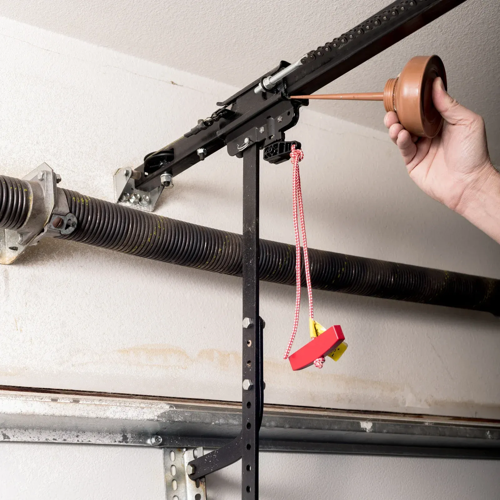 Person lubricating a garage door opener track with a brown oil can in a garage setting.