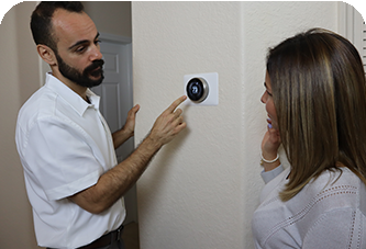Man points at smart thermostat on wall, explaining it to a woman. They are indoors.