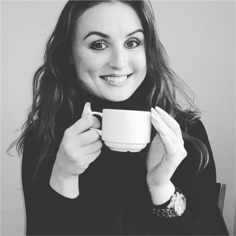 Woman with long dark hair, smiling, holding a white mug, wearing a black turtleneck, and watch.