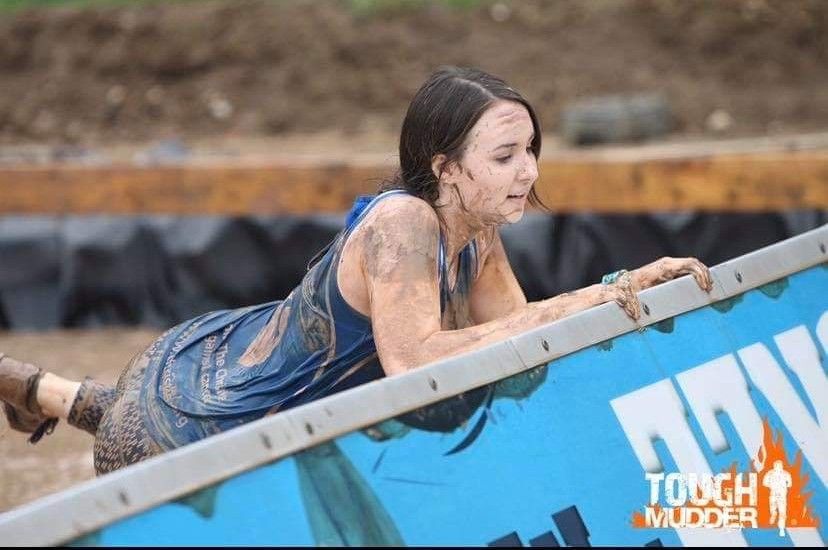 Woman covered in mud crawls under a blue obstacle during a Tough Mudder event.