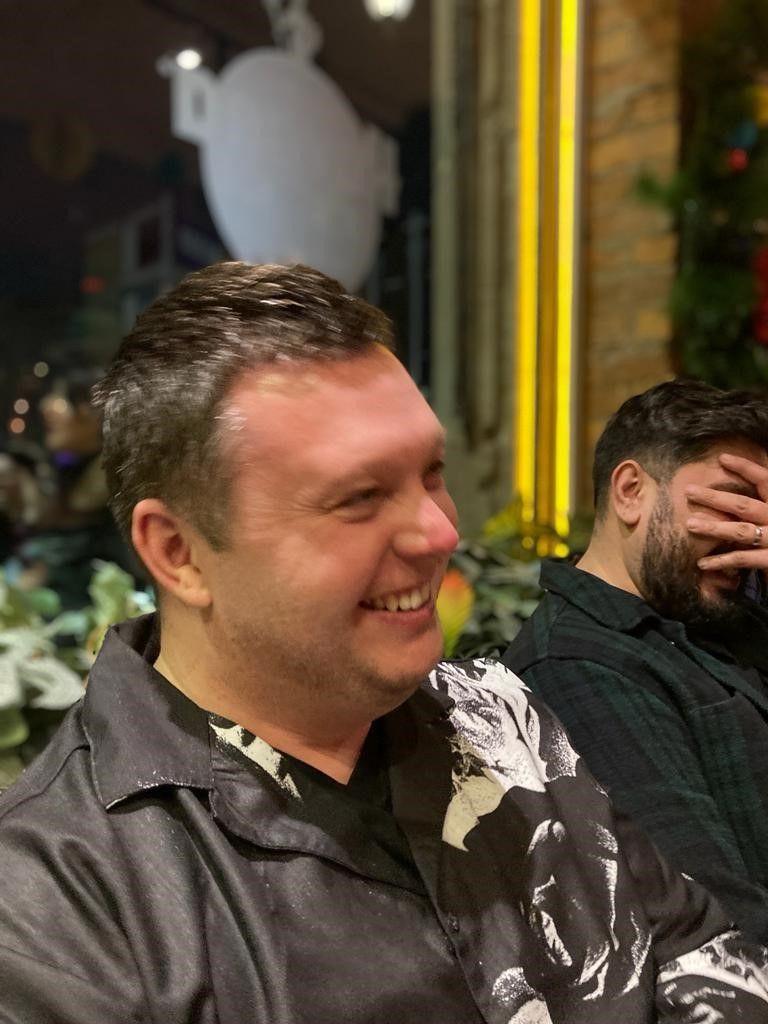 Man in black patterned shirt smiles, seated next to a person with hand over face, indoors.